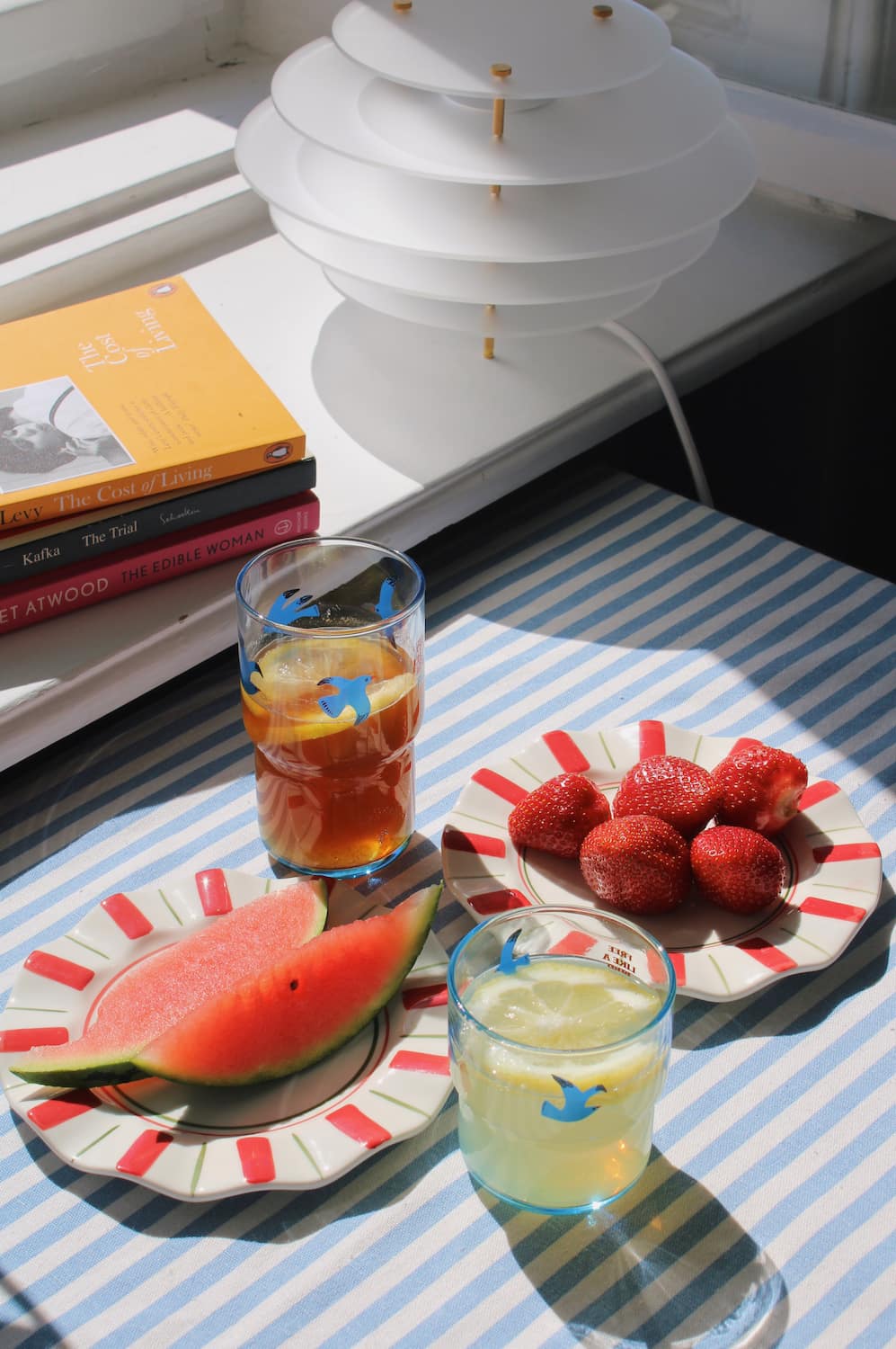 Two Riviera Ceramic Plates styled with watermelon and strawberries alongside the Birdie Tall Glass and Birdie Short Glass on a blue striped surface, with stacked books and a white pendant lamp in the background.