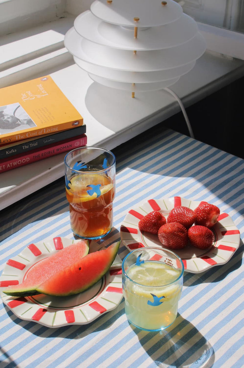 Two Riviera Ceramic Plates styled with watermelon and strawberries alongside the Birdie Tall Glass and Birdie Short Glass on a blue striped surface, with stacked books and a white pendant lamp in the background.