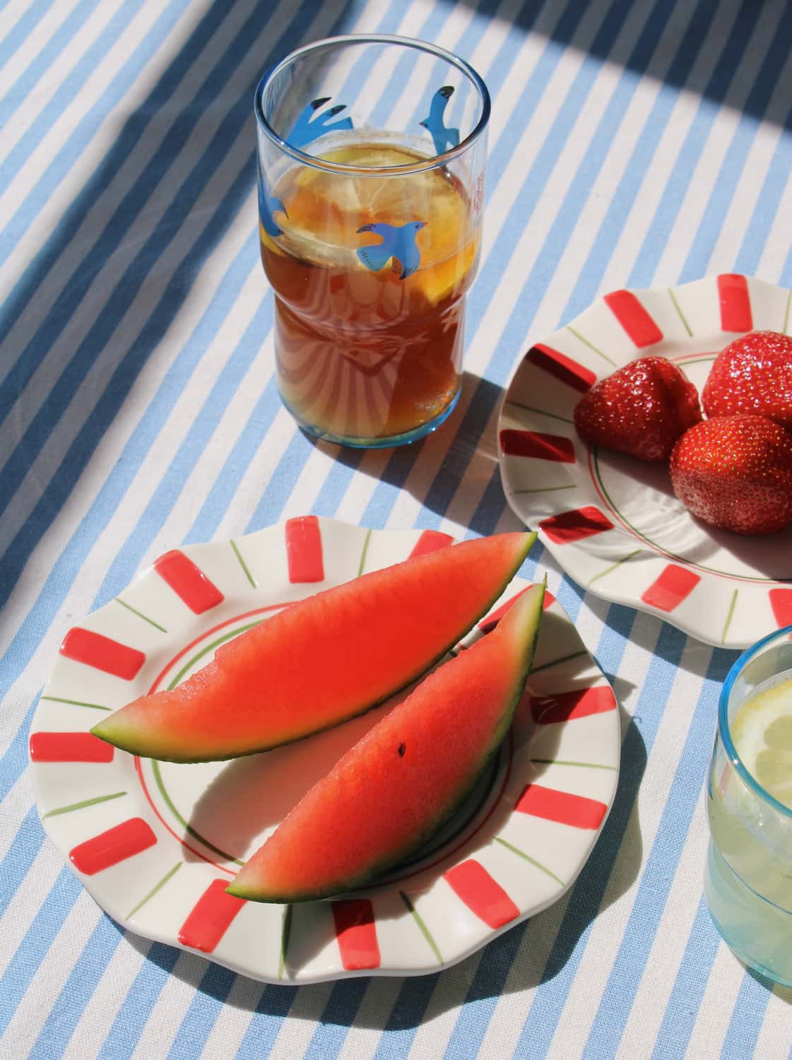 Two Riviera Ceramic Plates styled with watermelon and strawberries alongside the Birdie Tall Glass and Birdie Short Glass on a blue striped surface, with stacked books and a white pendant lamp in the background.