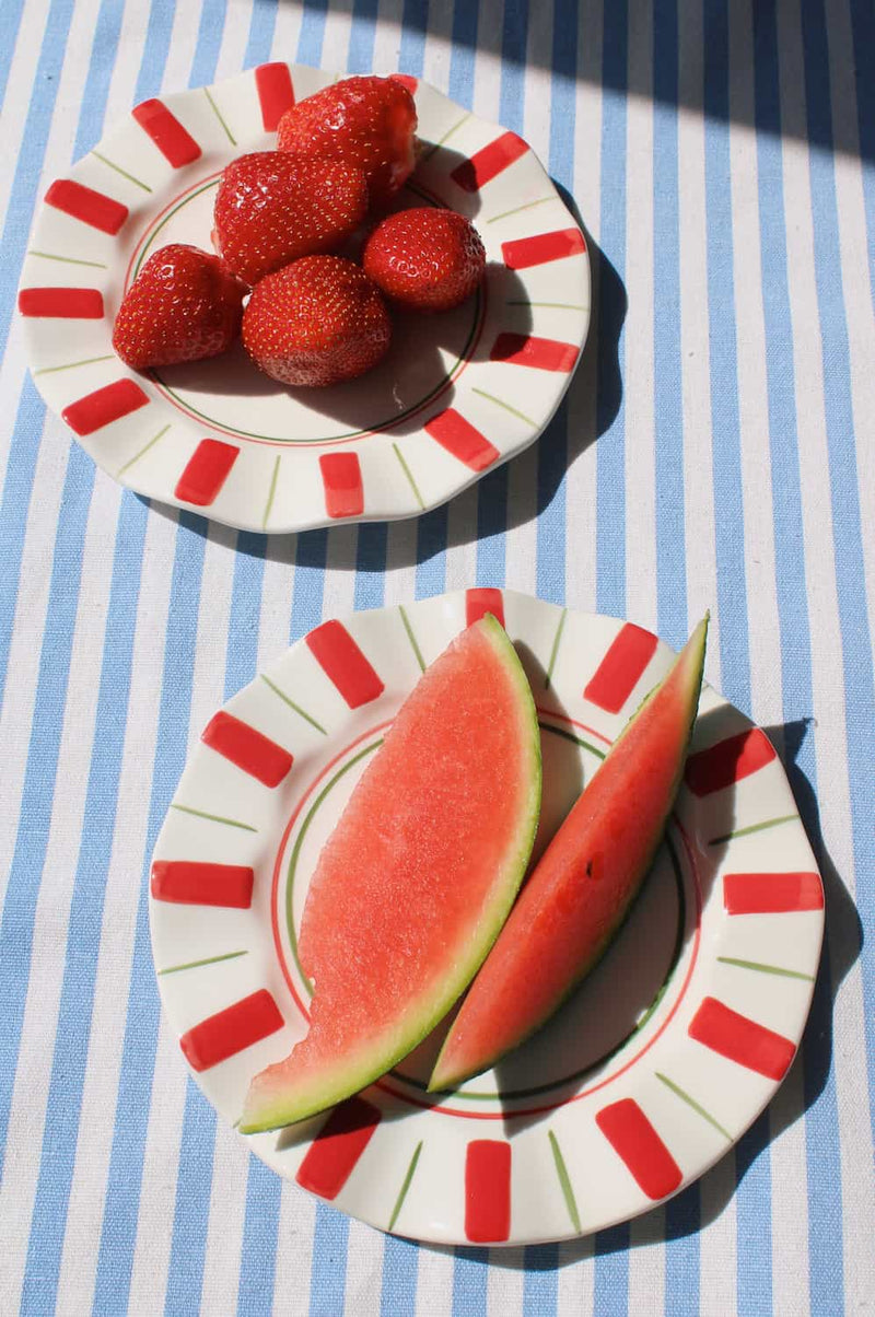 Two Riviera Ceramic Plates in cream with red stripe and green line border, styled with fresh strawberries and watermelon slices on a blue striped surface in natural sunlight.