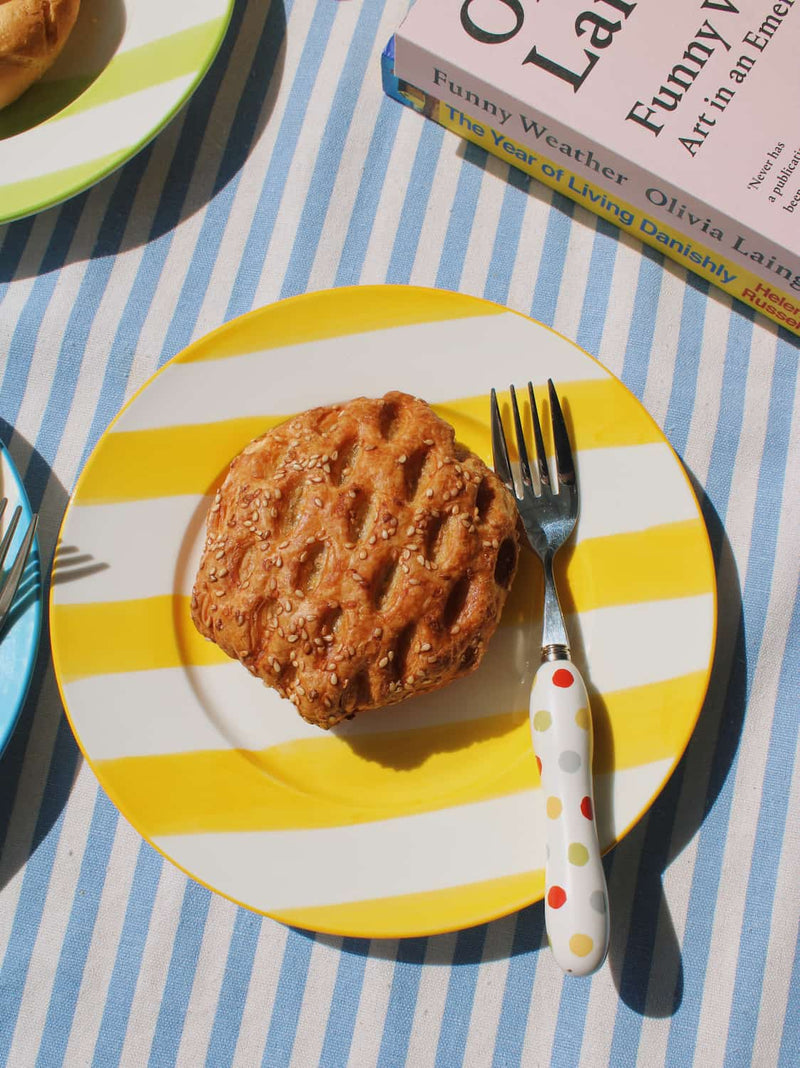 Etta Ceramic Plate in yellow with bold stripe pattern, styled with a sesame pastry and the Confetti Cutlery Set fork on a blue striped surface