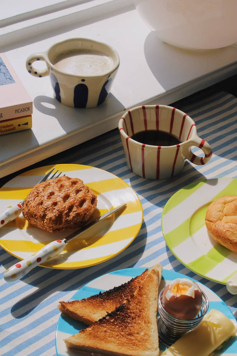 Etta Ceramic Plate in yellow with bold stripe pattern, styled with a sesame pastry and the Confetti Cutlery Set fork on a blue striped surface, with stacked books and the Olia Ceramic Plate visible in the background.