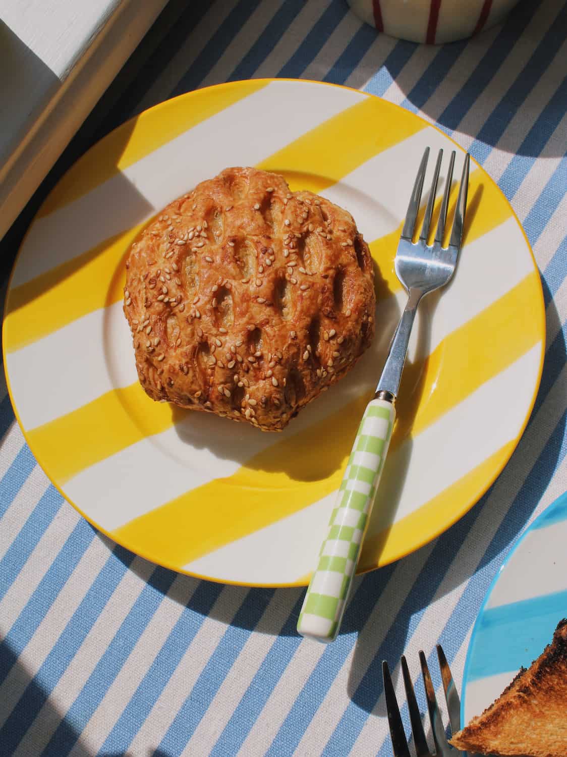 Etta Ceramic Plate in yellow with bold stripe pattern, styled with a sesame pastry and the Nico Cutlery Set fork on a blue striped surface