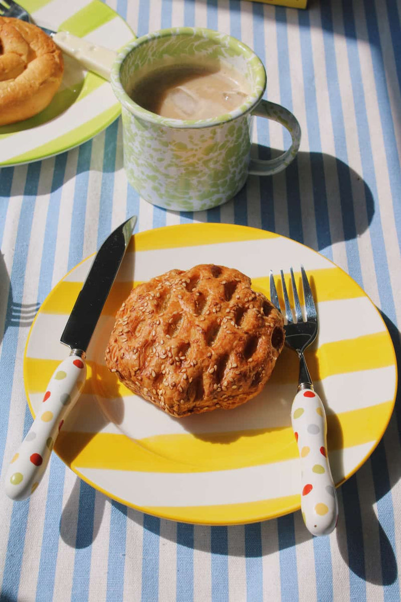Etta Ceramic Plate in yellow with bold stripe pattern, styled with a sesame pastry and the Confetti Cutlery Set fork on a blue striped surface