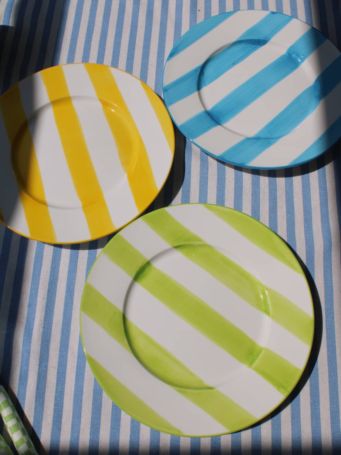 Etta Ceramic Plate in yellow, Mirel Ceramic Plate in blue and Olia Ceramic Plate in green, all with bold diagonal stripe patterns, displayed together on a blue striped surface in natural sunlight.