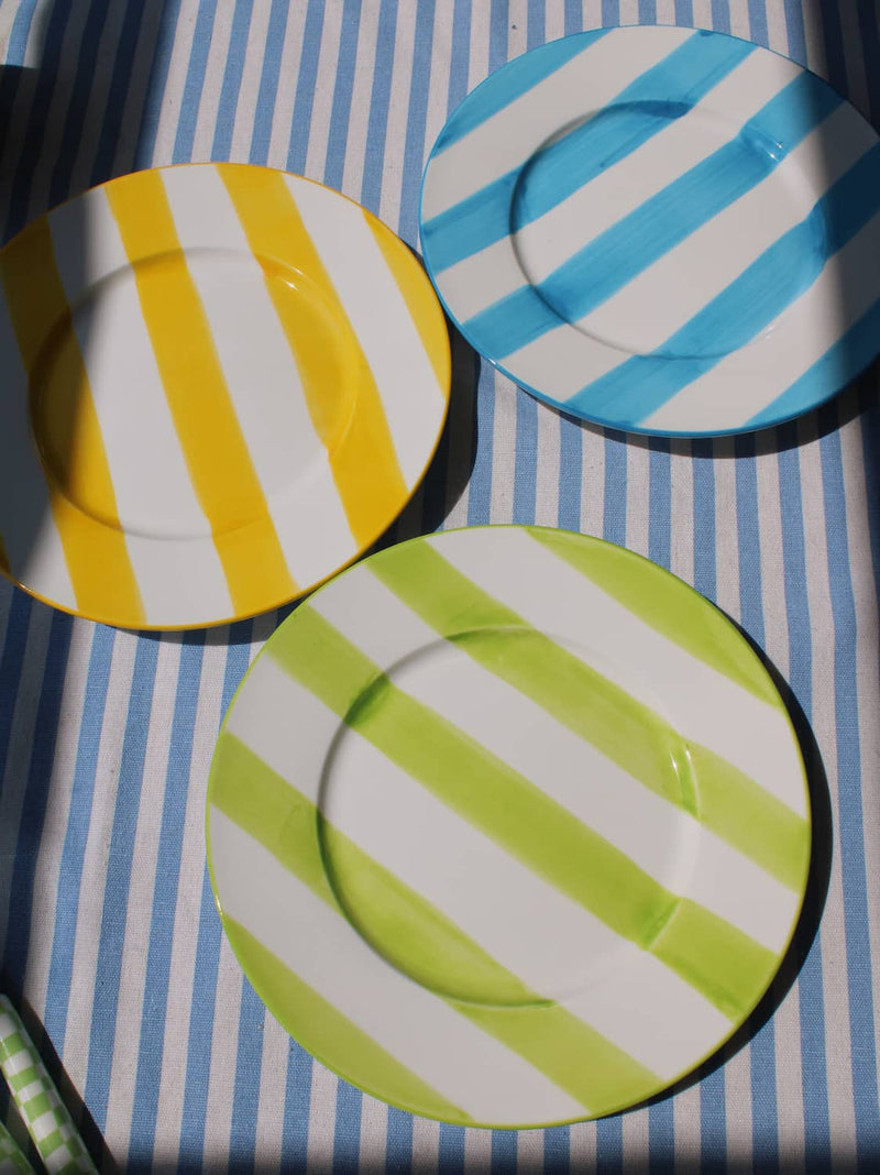 Etta Ceramic Plate in yellow, Mirel Ceramic Plate in blue and Olia Ceramic Plate in green, all with bold diagonal stripe patterns, displayed together on a blue striped surface in natural sunlight.