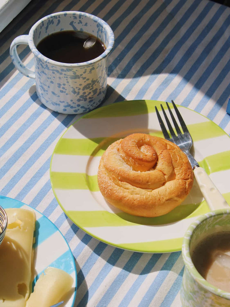 Olia Ceramic Plate in green with bold stripe pattern, styled with a cinnamon roll beside the Aeris Enamel Mug in blue speckle on a blue striped surface.