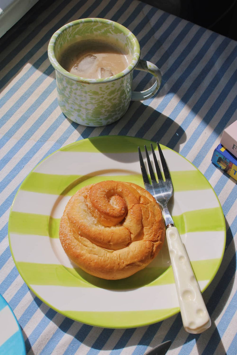 Olia Ceramic Plate in green with bold stripe pattern, styled with a cinnamon roll beside the Clover Enamel Mug in green speckle on a blue striped surface.