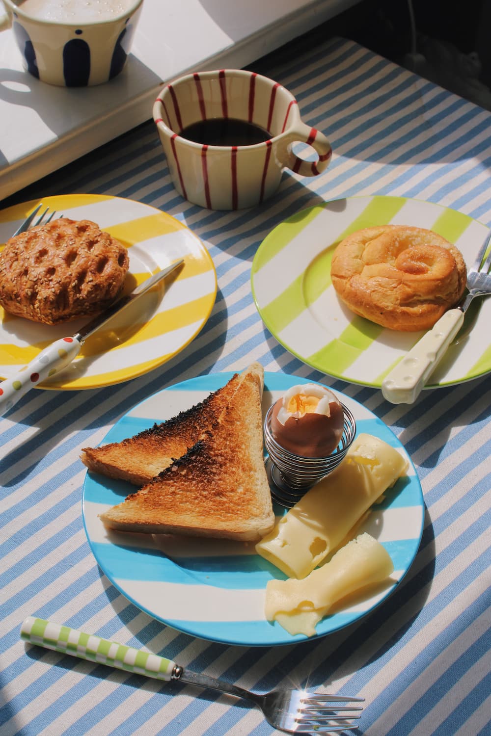 Olia Ceramic Plate, Etta Ceramic Plate and Mirel Ceramic Plate styled with a cinnamon roll, sesame pastry and soft boiled egg breakfast spread, alongside the Arlo Ceramic Mug and Enzo Ceramic Mug on a blue striped surface with the Nico Cutlery Set.