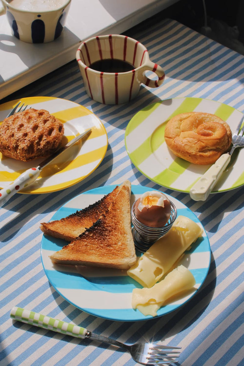 Olia Ceramic Plate, Etta Ceramic Plate and Mirel Ceramic Plate styled with a cinnamon roll, sesame pastry and soft boiled egg breakfast spread, alongside the Arlo Ceramic Mug and Enzo Ceramic Mug on a blue striped surface with the Nico Cutlery Set.