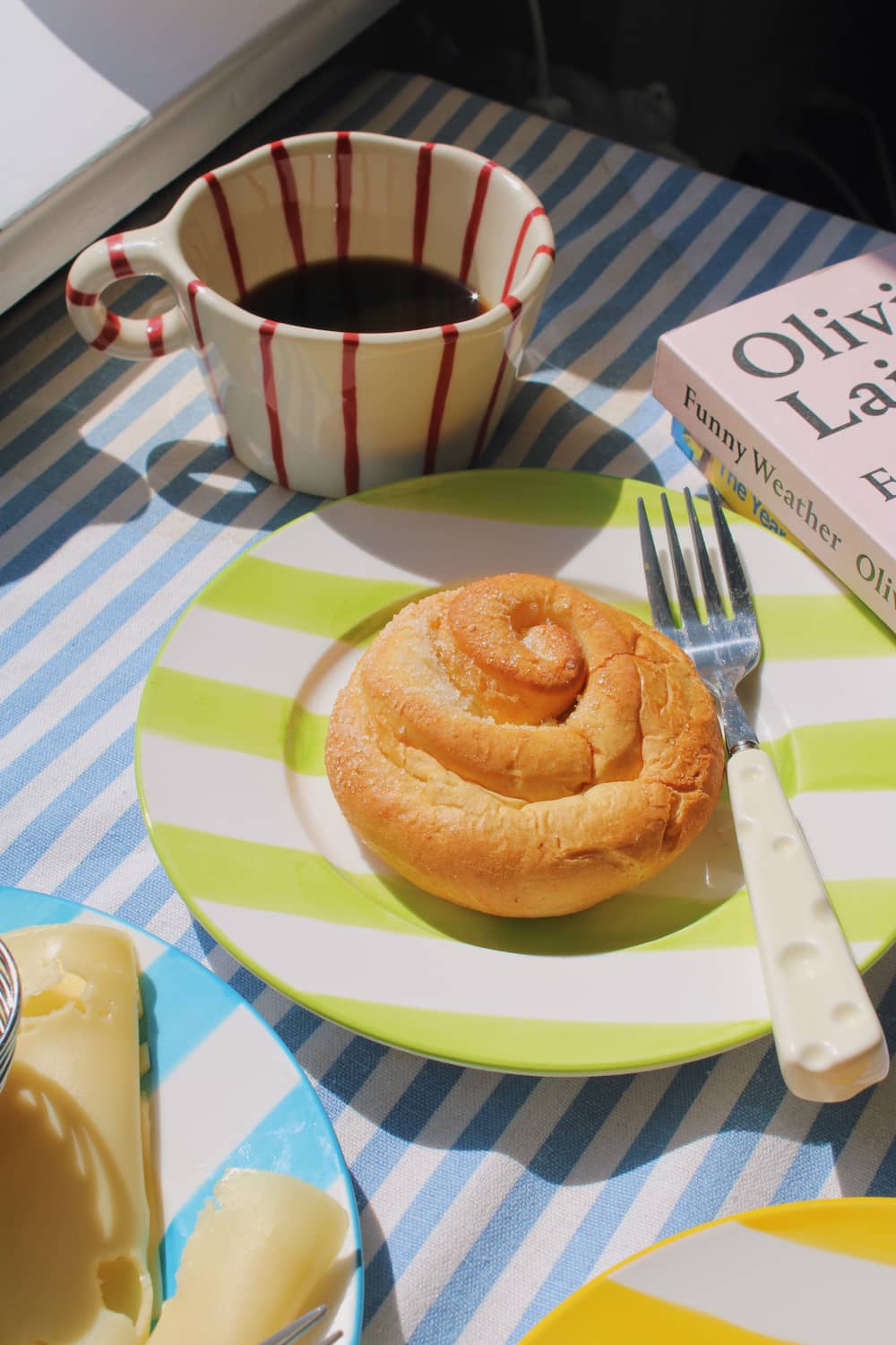 Olia Ceramic Plate in green with bold stripe pattern, styled with a cinnamon roll beside the Arlo Ceramic Mug in cream with red stripes, with stacked books and the Mirel Ceramic Plate visible in the background.
