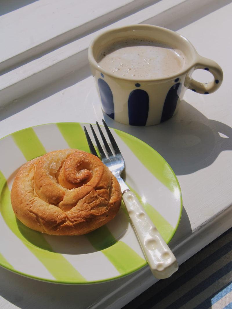 Olia Ceramic Plate in green with bold stripe pattern, styled with a cinnamon roll beside the Enzo Ceramic Mug on a white windowsill in natural sunlight.