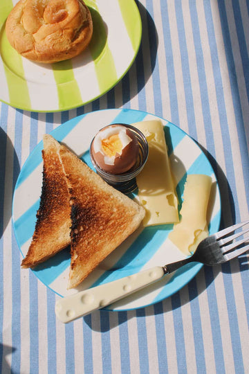 Mirel Ceramic Plate in white with bold blue stripe pattern, styled with toast, a soft boiled egg and cheese slices on a blue striped surface.