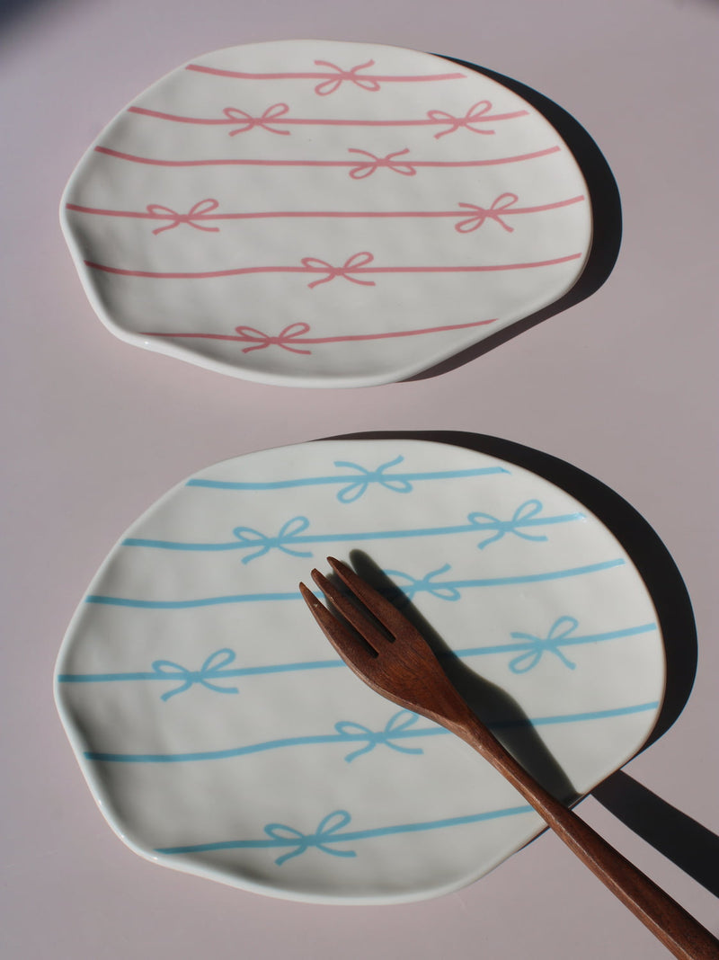 Rina and Lina Ceramic Plates side by side, blue and pink bow ribbon pattern, styled with a wooden fork, handmade ceramic plates