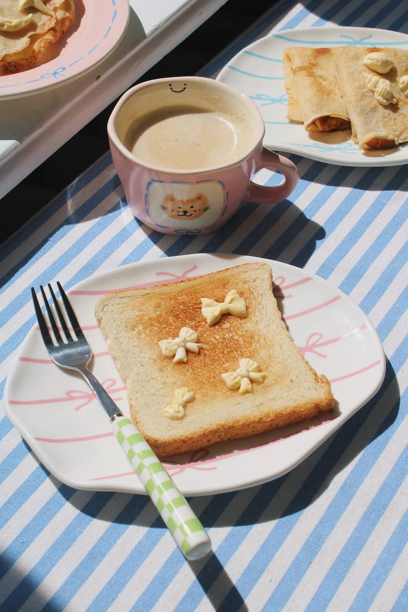Lina Ceramic Plate in white with pink bow motifs, styled with buttered toast and a green gingham fork beside the Mimi Ceramic Mug on a blue striped surface.