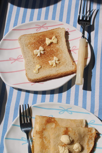 Lina Ceramic Plate and Rina Ceramic Plate side by side — pink and blue bow motifs — styled with buttered toast and crepes on a blue striped surface in natural sunlight.