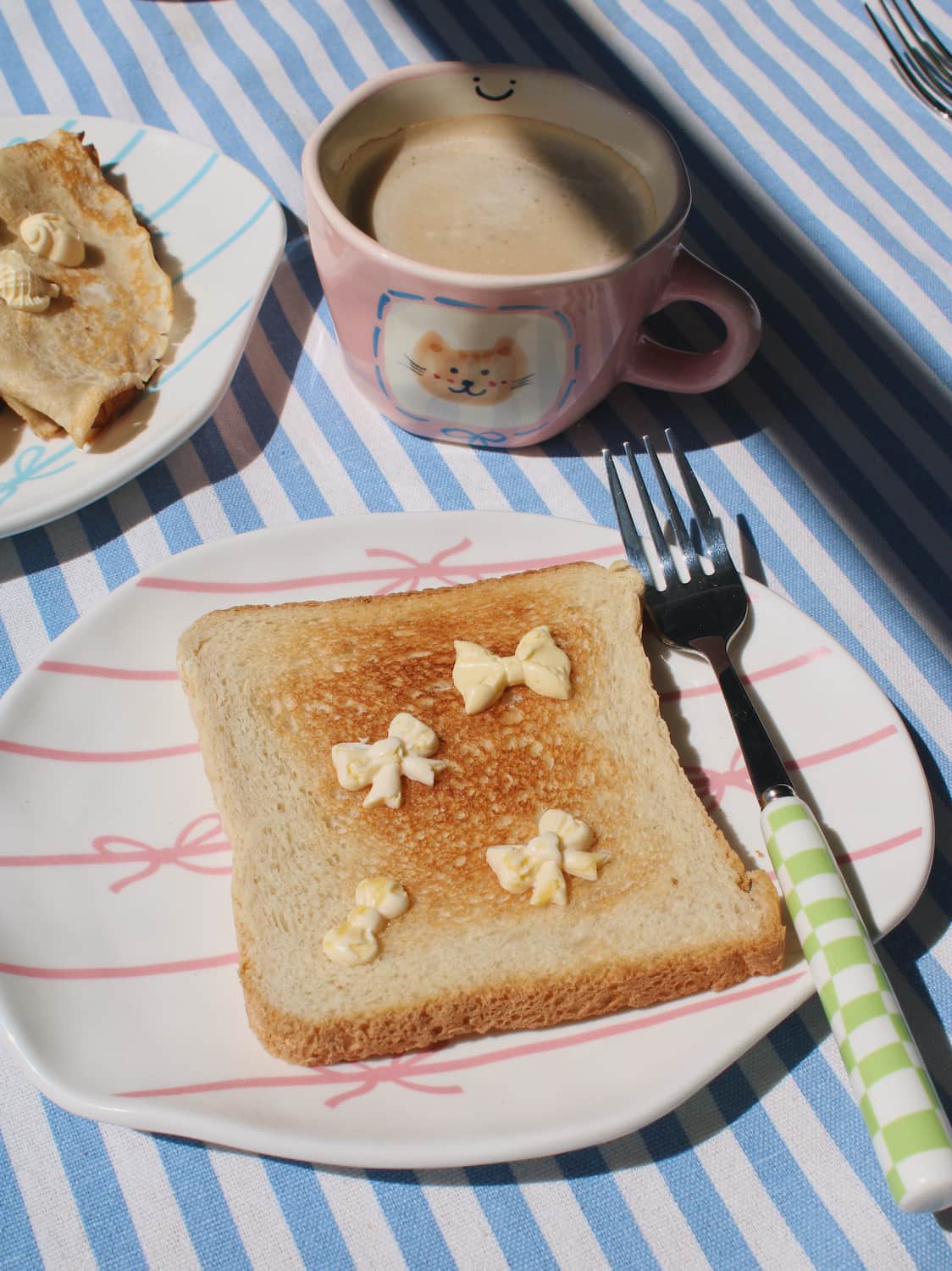 Lina Ceramic Plate in white with pink bow motifs, styled with buttered toast and a green gingham fork beside the Mimi Ceramic Mug on a blue striped surface.