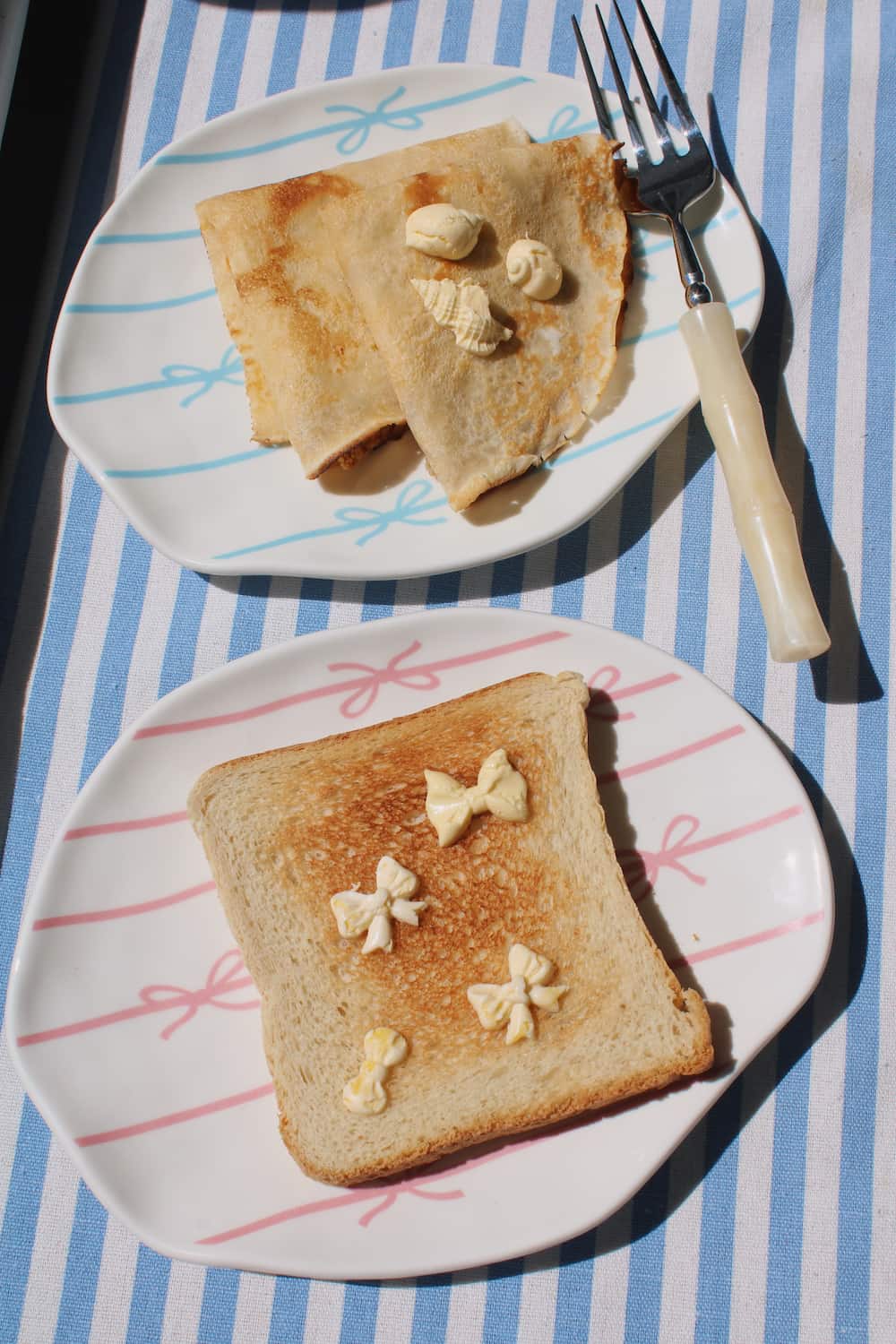 Lina Ceramic Plate and Rina Ceramic Plate side by side — pink and blue bow motifs — styled with buttered toast and crepes on a blue striped surface in natural sunlight.