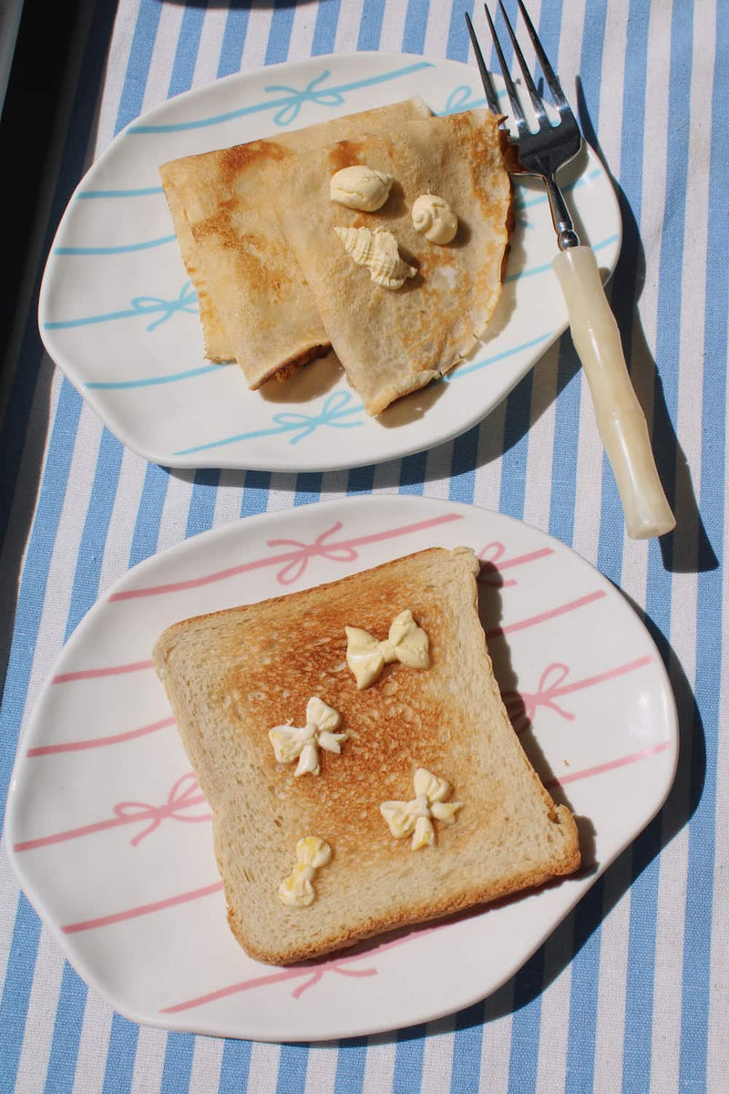 Lina Ceramic Plate and Rina Ceramic Plate side by side — pink and blue bow motifs — styled with buttered toast and crepes on a blue striped surface in natural sunlight.