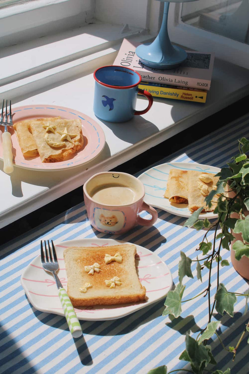 Lina Ceramic Plate and Rina Ceramic Plate styled with buttered toast and crepes alongside the Mimi Ceramic Mug and Birdie Ceramic Mug, on a blue striped surface in natural sunlight.