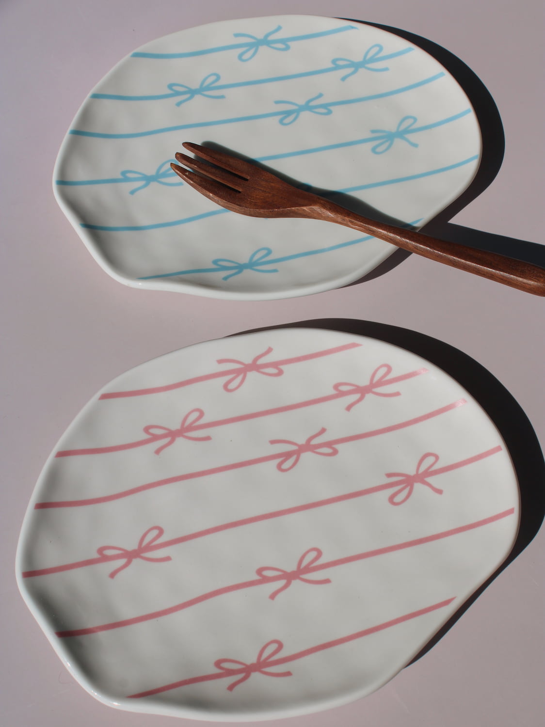Rina and Lina Ceramic Plates side by side, blue and pink bow ribbon pattern, styled with a wooden fork, handmade ceramic plates