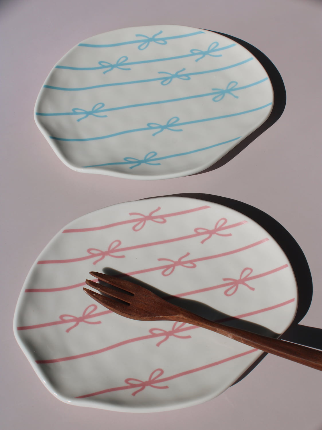 Rina and Lina Ceramic Plates side by side, blue and pink bow ribbon pattern, styled with a wooden fork, handmade ceramic plates