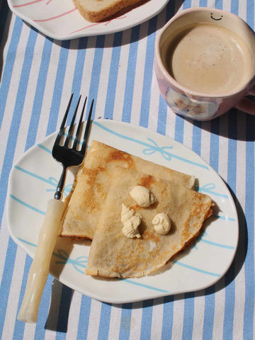 Rina Ceramic Plate in white with blue bow motifs, styled with buttered crepes and a fork on a blue striped surface, with the Mimi Ceramic Mug and Lina Ceramic Plate visible in the background.