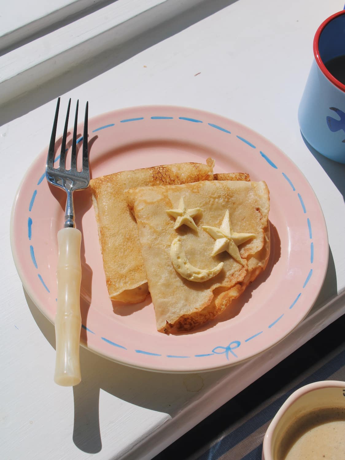 Mimi Ceramic Plate in pink with blue dashed border and bow motif, styled with a buttered crepe on a blue striped surface, with the Mimi Ceramic Mug visible in the background.