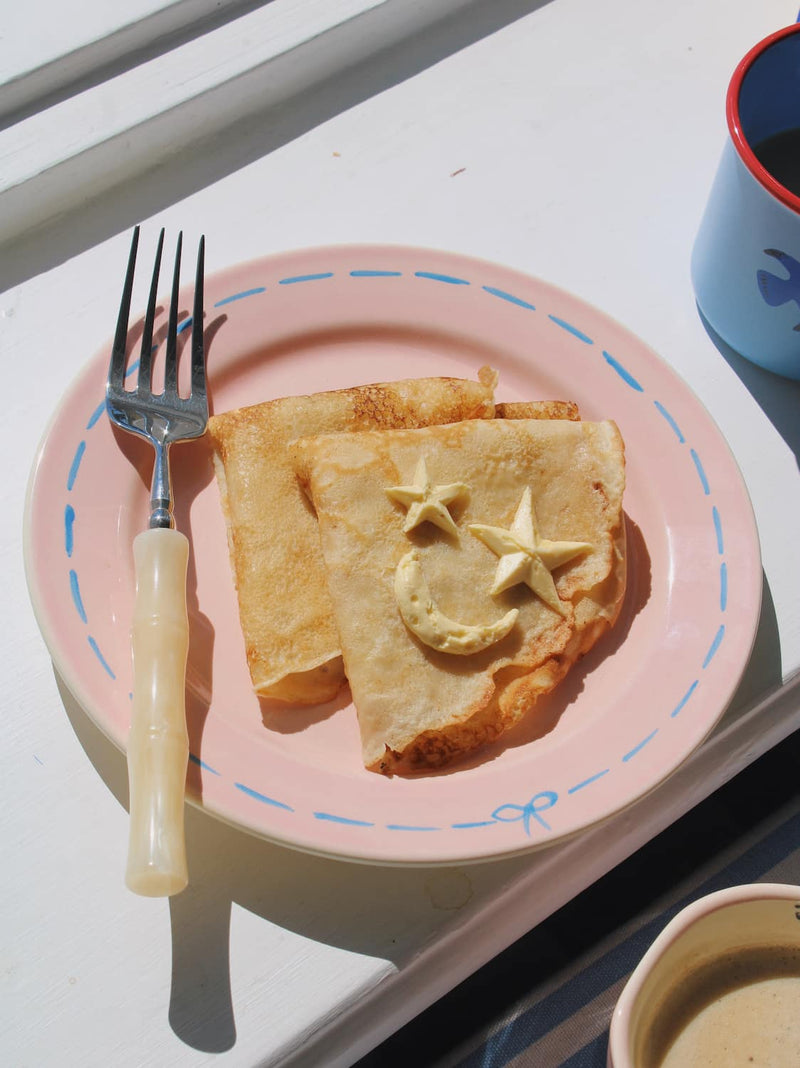 Mimi Ceramic Plate in pink with blue dashed border and bow motif, styled with a buttered crepe on a blue striped surface, with the Mimi Ceramic Mug visible in the background.