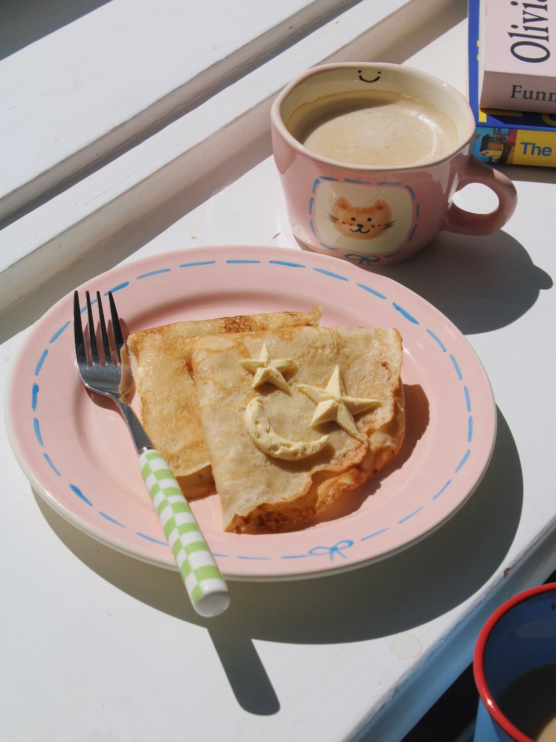 Mimi Ceramic Mug filled with coffee beside the Mimi Ceramic Plate styled with a buttered crepe on a blue striped surface, with ivy in the background.