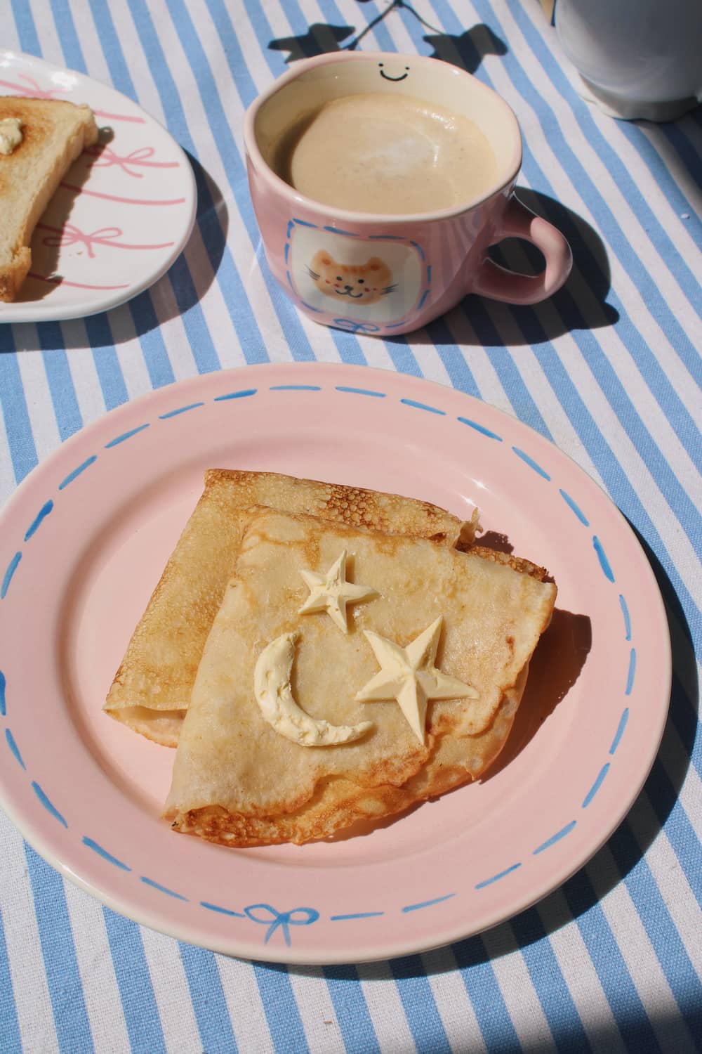 Mimi Ceramic Mug filled with coffee beside the Mimi Ceramic Plate styled with a buttered crepe on a blue striped surface, with ivy in the background.