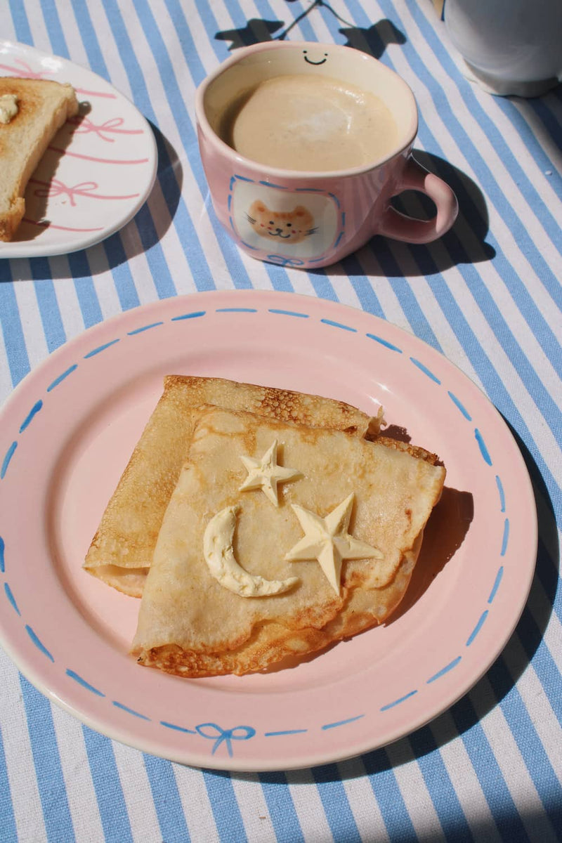 Mimi Ceramic Mug filled with coffee beside the Mimi Ceramic Plate styled with a buttered crepe on a blue striped surface, with ivy in the background.