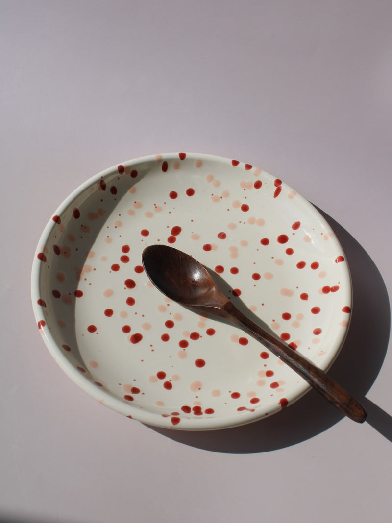 Bera Ceramic Plate in cream with red speckled dot pattern, paired with a wooden fork, viewed from above on a soft pink background.