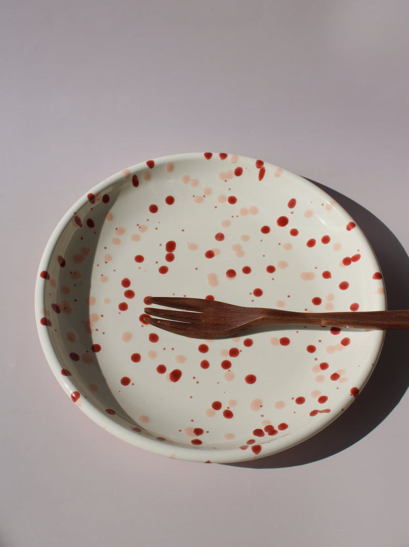 Bera Ceramic Plate in cream with red speckled dot pattern, paired with a wooden fork, viewed from above on a soft pink background.