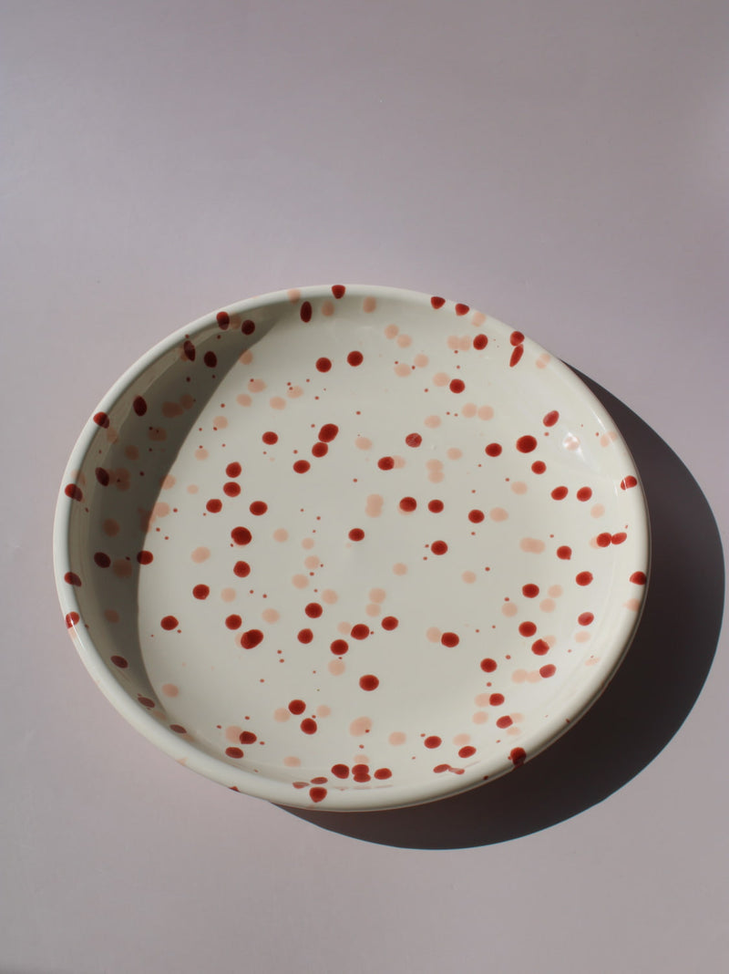 Bera Ceramic Plate in cream with red speckled dot pattern, paired with a wooden fork, viewed from above on a soft pink background.