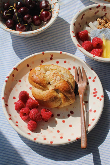 Bera Ceramic Plate in cream with red speckled dot pattern, styled with an almond pastry and fresh raspberries with a wooden fork, with the Naro Ceramic Bowl filled with cherries and the Bera Ceramic Bowl with granola visible in the background on a blue striped surface.