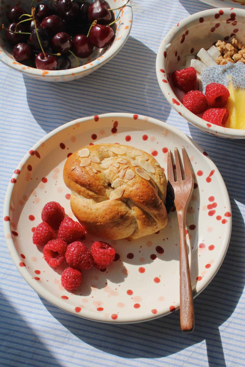 Bera Ceramic Plate in cream with red speckled dot pattern, styled with an almond pastry and fresh raspberries with a wooden fork, with the Naro Ceramic Bowl filled with cherries and the Bera Ceramic Bowl with granola visible in the background on a blue striped surface.