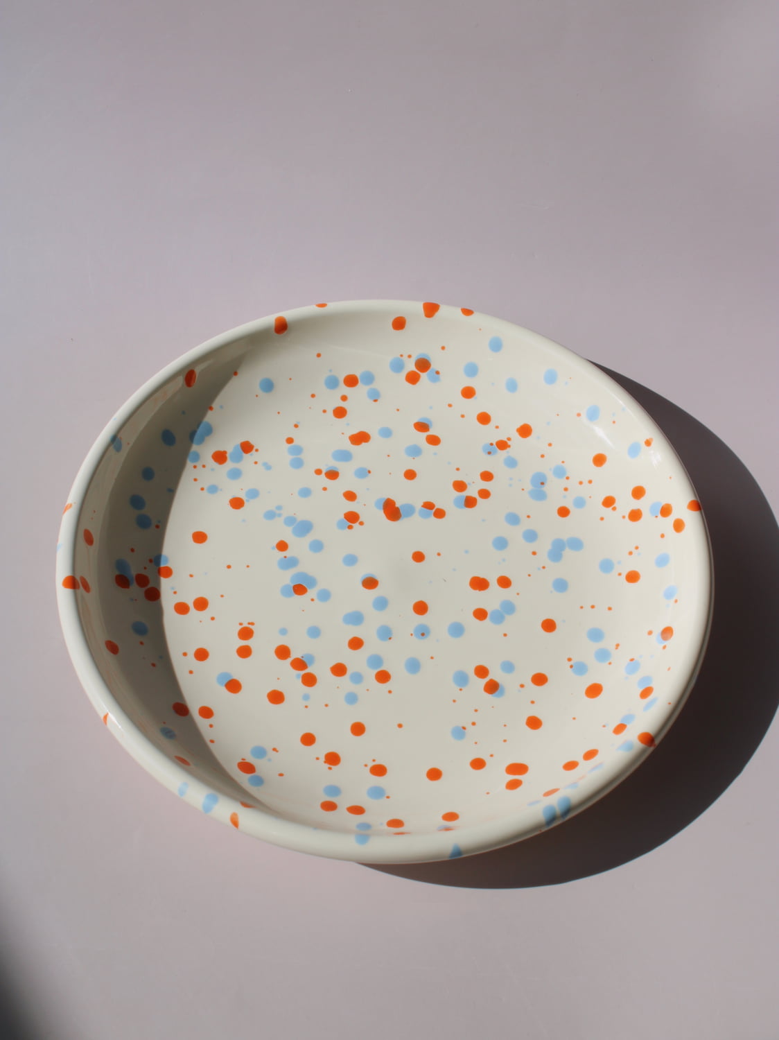 Naro Ceramic Plate in cream with orange and blue speckled dot pattern, viewed from above on a soft pink background.