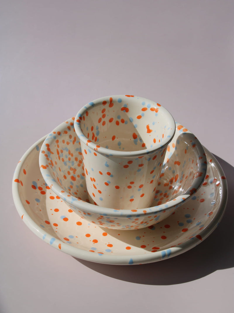 Naro Ceramic Cup, Bowl, and Plate in cream with orange and blue speckled dot pattern, on a soft pink background.