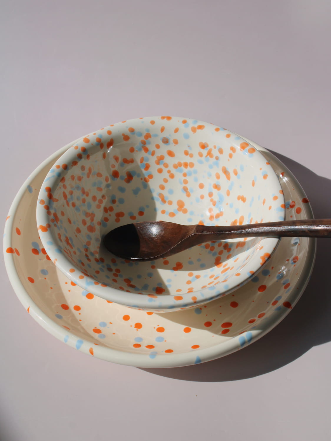 Naro Ceramic Cup, Bowl, and Plate in cream with orange and blue speckled dot pattern, on a soft pink background.