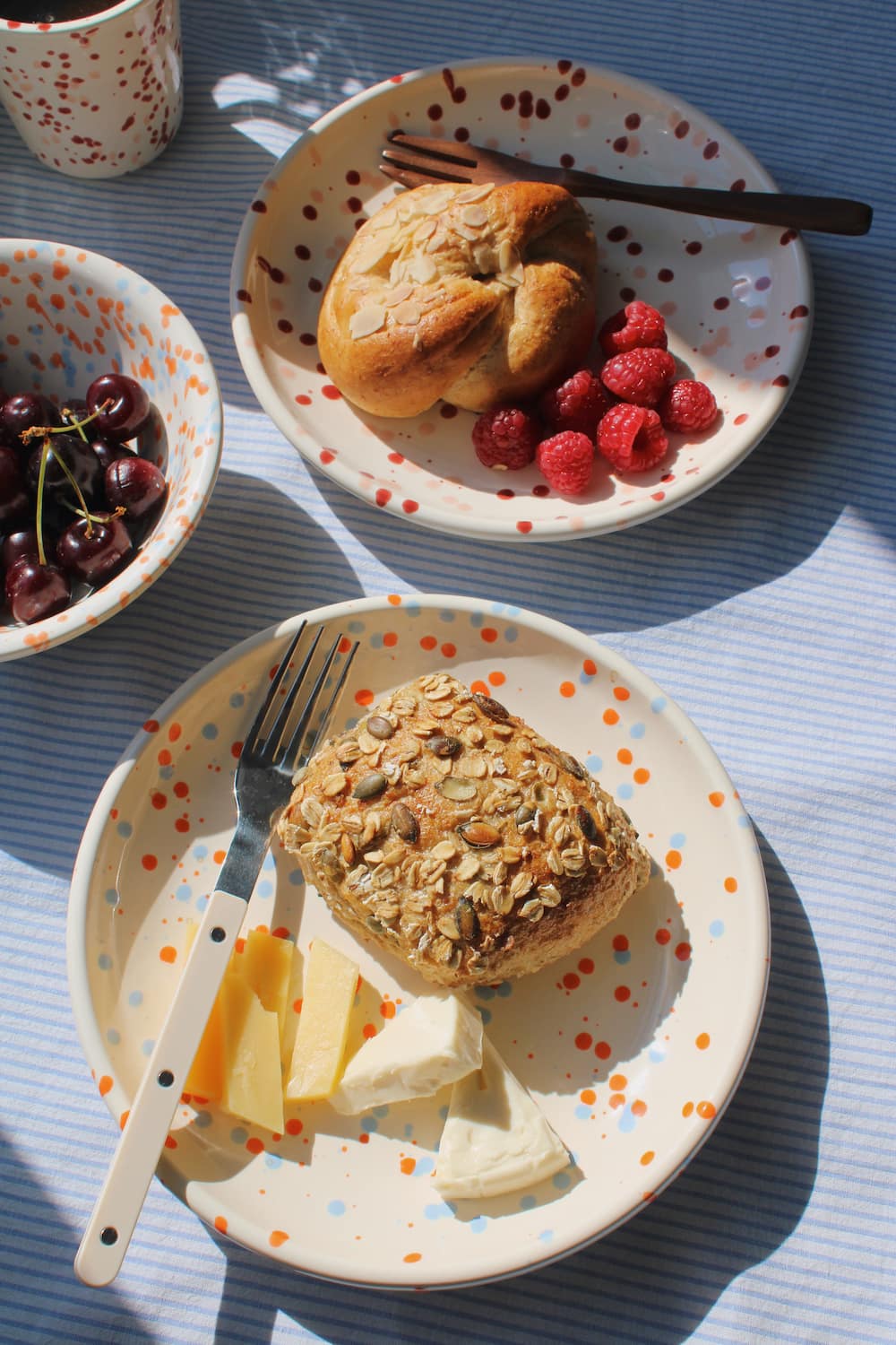 Naro Ceramic Plate styled with a seeded bread roll and cheese, with the Naro Ceramic Bowl filled with cherries and the Bera Ceramic Cup visible in the background on a blue striped surface in natural sunlight.