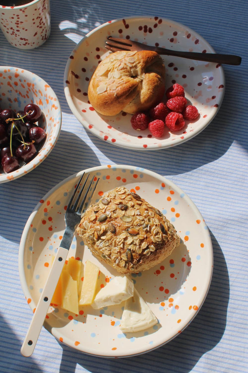Naro Ceramic Plate styled with a seeded bread roll and cheese, with the Naro Ceramic Bowl filled with cherries and the Bera Ceramic Cup visible in the background on a blue striped surface in natural sunlight.