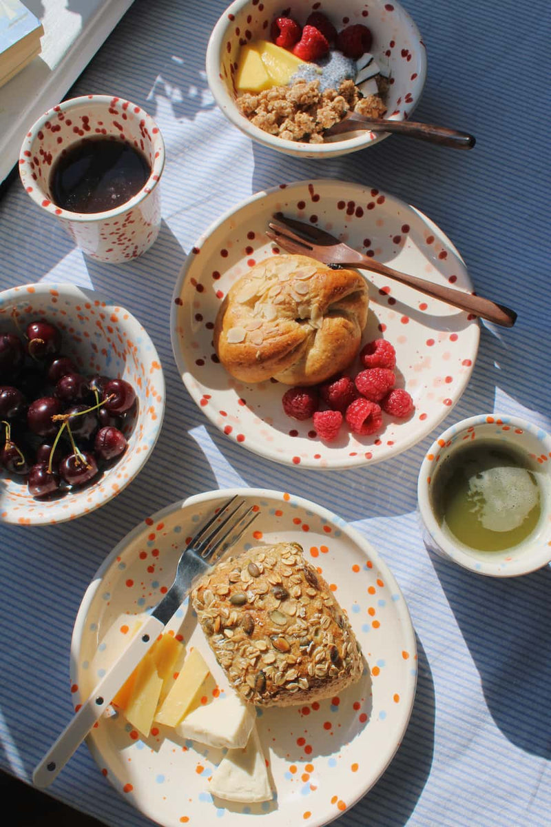 Naro Ceramic Plate, Bera Ceramic Plate, Naro Ceramic Bowl, Bera Ceramic Bowl, Naro Ceramic Cup and Bera Ceramic Cup styled together as a complete breakfast spread with pastries, cherries, granola and raspberries on a blue striped surface, with ivy and stacked books by a sunny window.