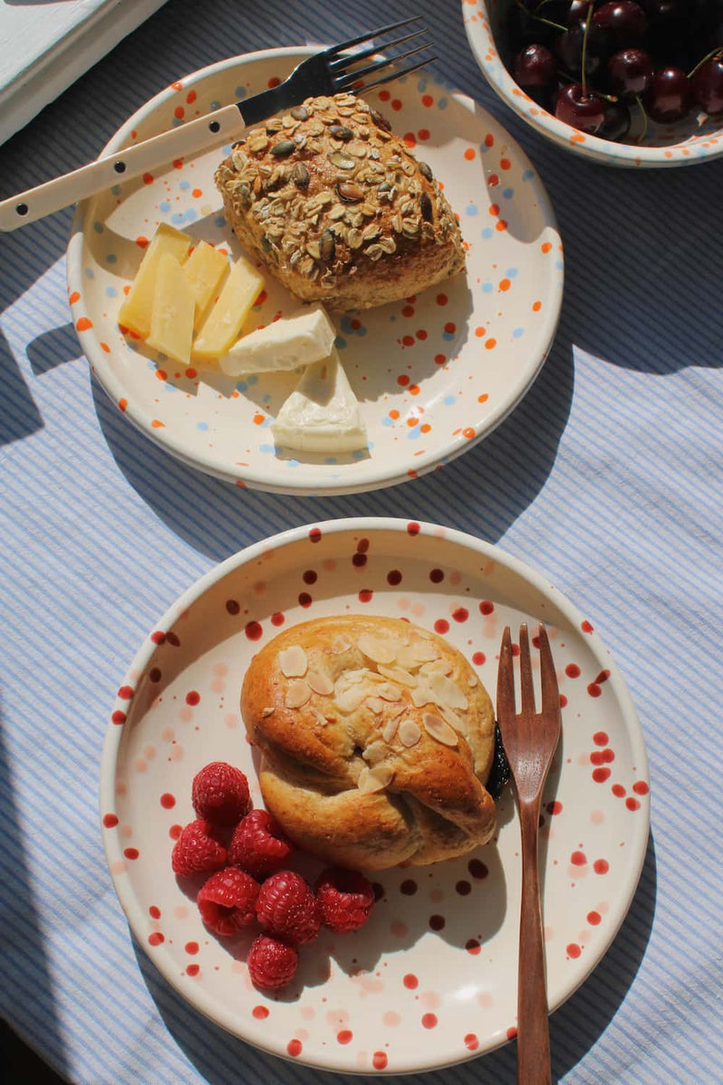 Naro Ceramic Plate with seeded bread roll and cheese alongside the Bera Ceramic Plate with almond pastry and raspberries, with the Naro Ceramic Bowl filled with cherries on a blue striped surface.