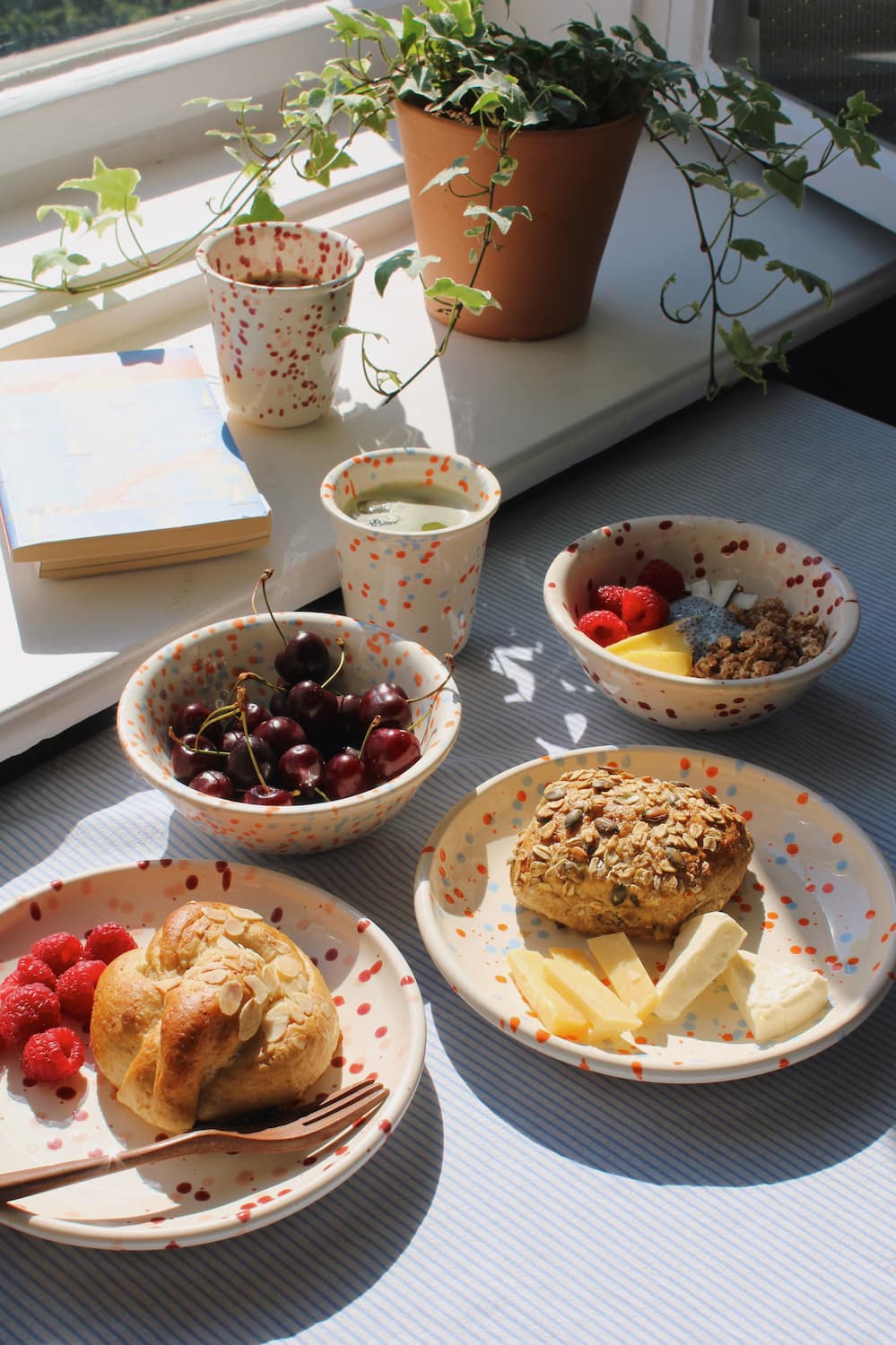 Naro Ceramic Plate, Bera Ceramic Plate, Naro Ceramic Bowl, Bera Ceramic Bowl, Naro Ceramic Cup and Bera Ceramic Cup styled together as a complete breakfast spread with pastries, cherries, granola and raspberries on a blue striped surface, with ivy and stacked books by a sunny window.