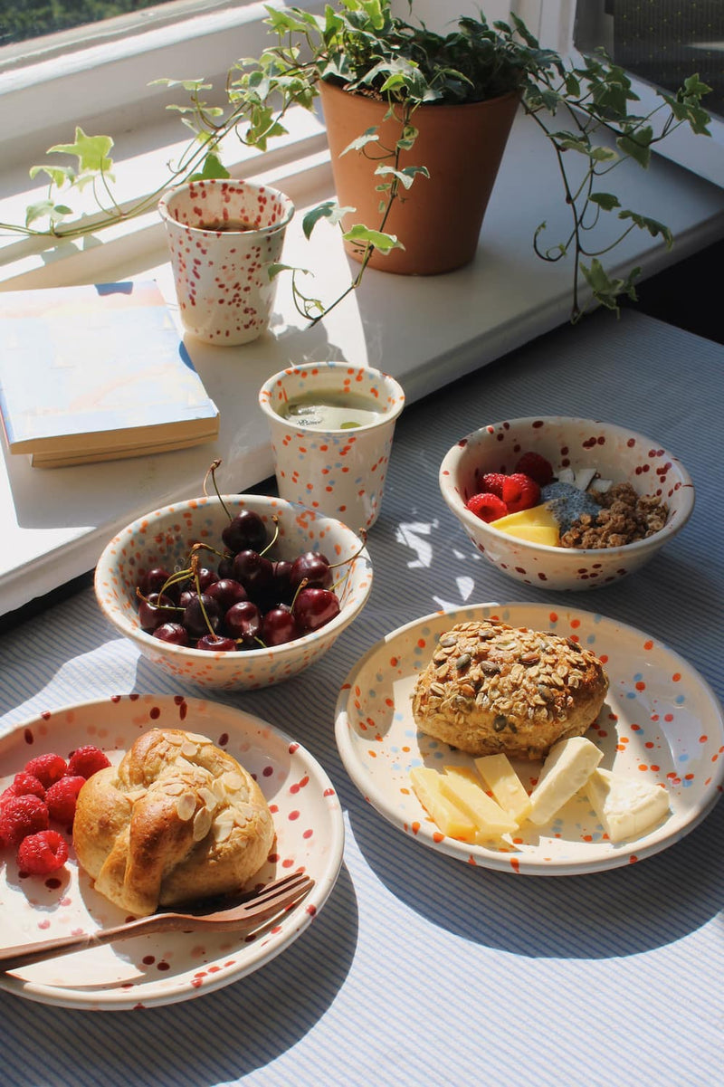 Naro Ceramic Plate, Bera Ceramic Plate, Naro Ceramic Bowl, Bera Ceramic Bowl, Naro Ceramic Cup and Bera Ceramic Cup styled together as a complete breakfast spread with pastries, cherries, granola and raspberries on a blue striped surface, with ivy and stacked books by a sunny window.