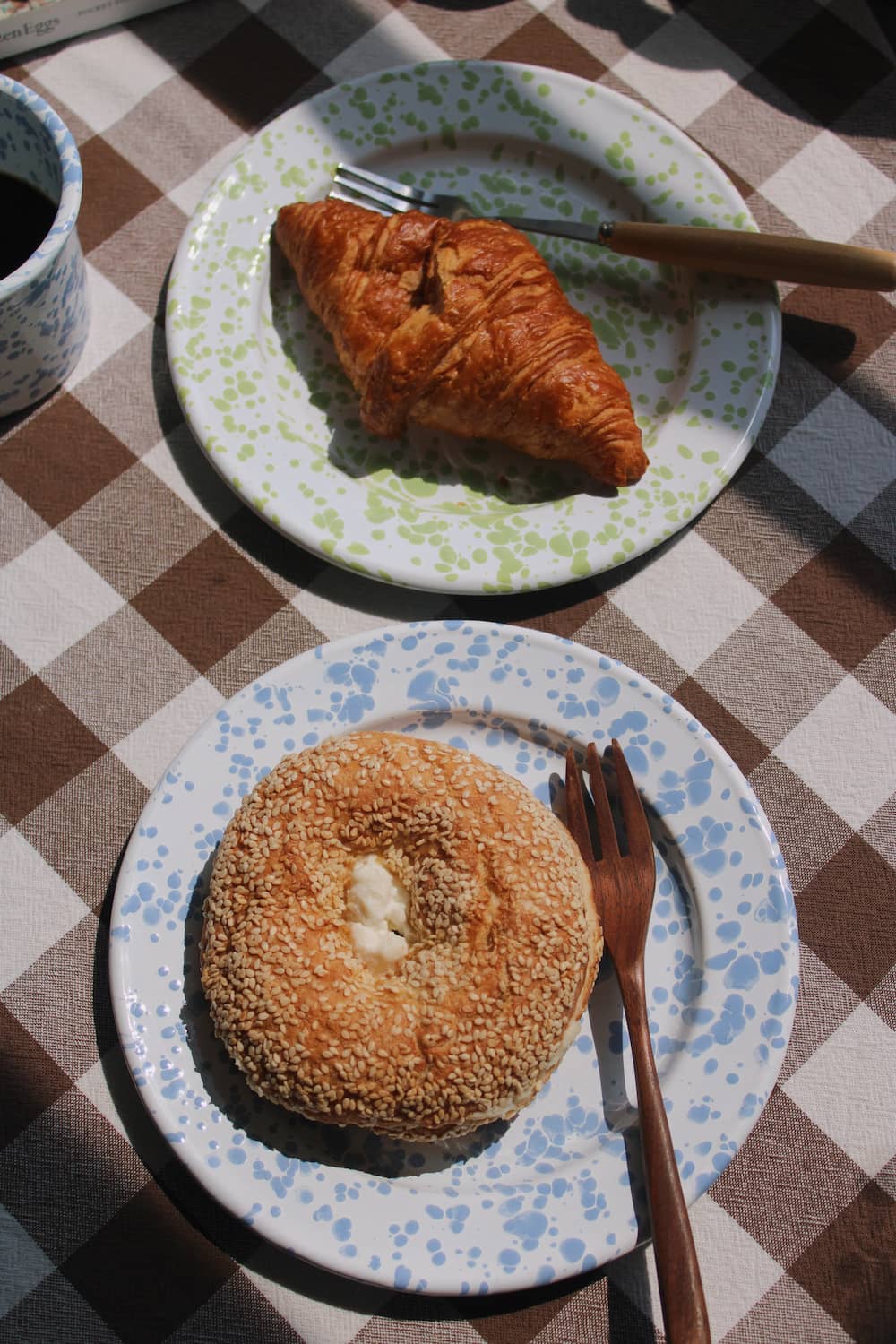 Aeris Enamel Plate in blue speckle with a sesame bagel and wooden fork alongside the Clover Enamel Plate in green speckle with a croissant, on a brown gingham surface with the Aeris Enamel Mug visible in the background.