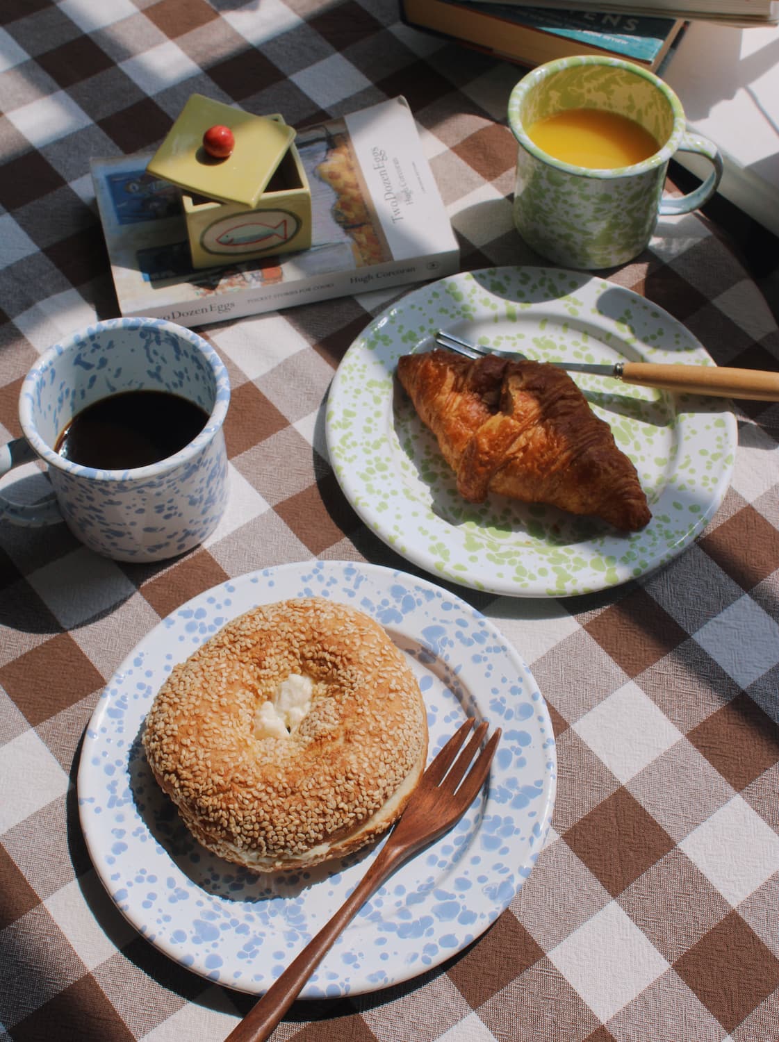 Aeris Enamel Mug and Aeris Enamel Plate styled with a sesame bagel alongside the Clover Enamel Mug and Clover Enamel Plate with a croissant, on a brown gingham surface with stacked books, flowers and a terracotta pot by a sunny window.