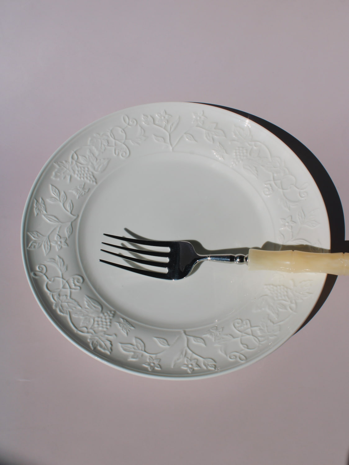 Florentia Ceramic Plate in white with embossed floral and botanical border detail, styled with a silver fork, on a soft pink background.