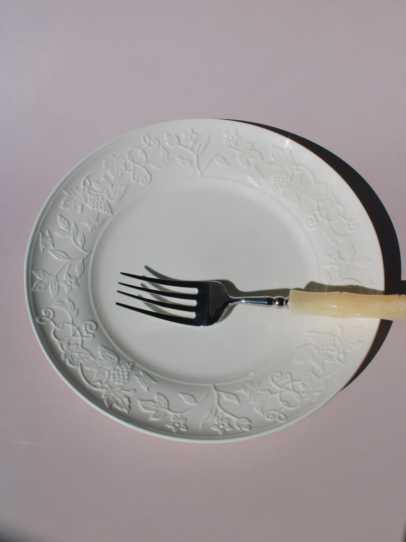 Florentia Ceramic Plate in white with embossed floral and botanical border detail, styled with a silver fork, on a soft pink background.